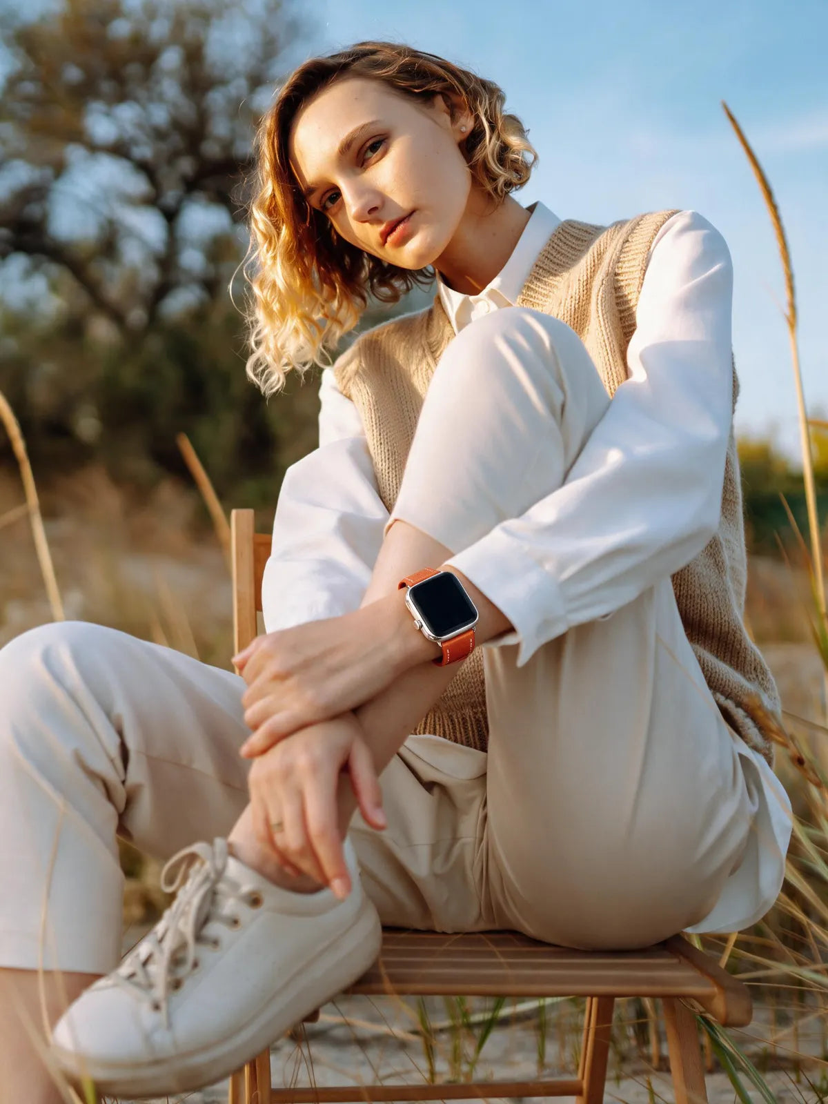 A woman sits on a wooden chair outdoors wearing a beige knit sweater vest over a white collared shirt, cream pants, and white sneakers. She is wearing a smartwatch with a tan strap and a black watch face.