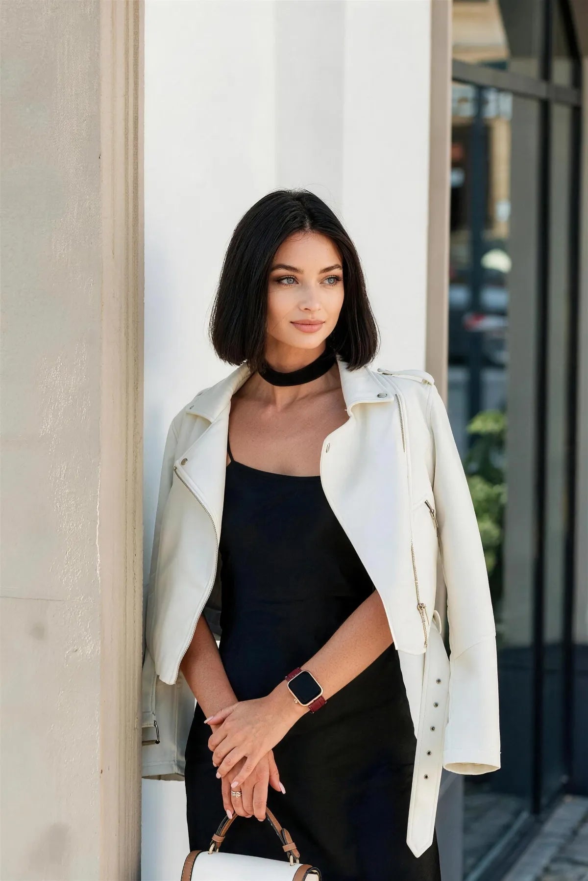 A woman with a short dark bob stands outdoors wearing a black dress, a cream leather moto jacket, and a black choker. She is holding a small white and brown handbag.