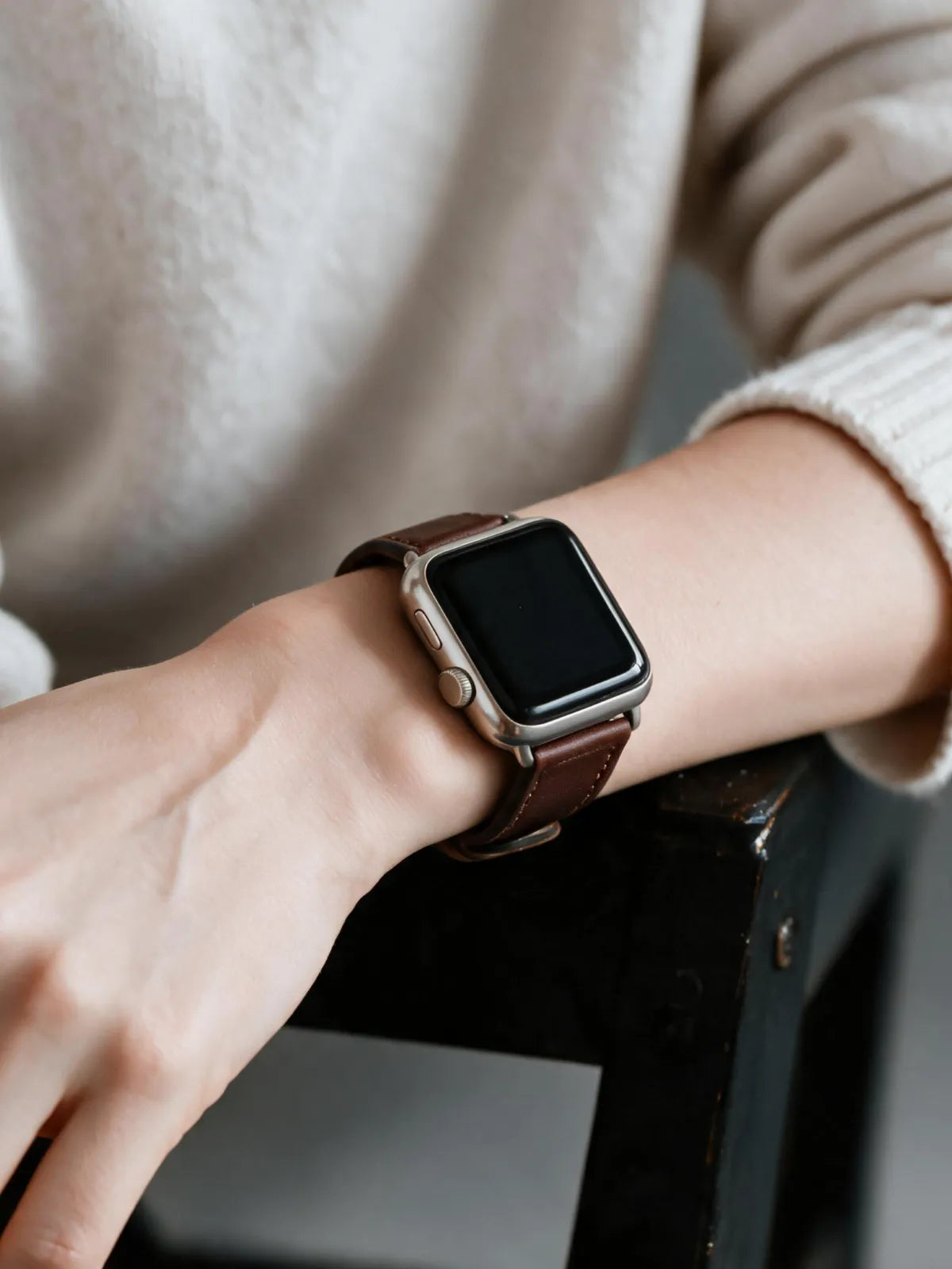 A close-up of a person's wrist wearing a smartwatch with a brown leather strap and a silver-toned metal case, set against a soft beige knit sweater.
