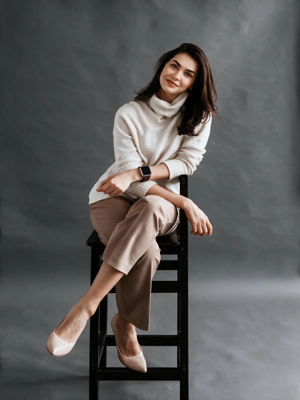 A woman sits on a black stool against a dark gray studio background, wearing a cream turtleneck sweater, beige cropped pants, and beige pointed flats.