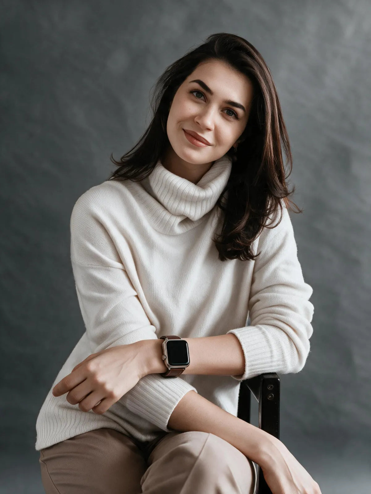A woman with dark hair sits wearing a cream ribbed turtleneck sweater and beige pants, with a smartwatch on her wrist, against a dark gray textured studio background.