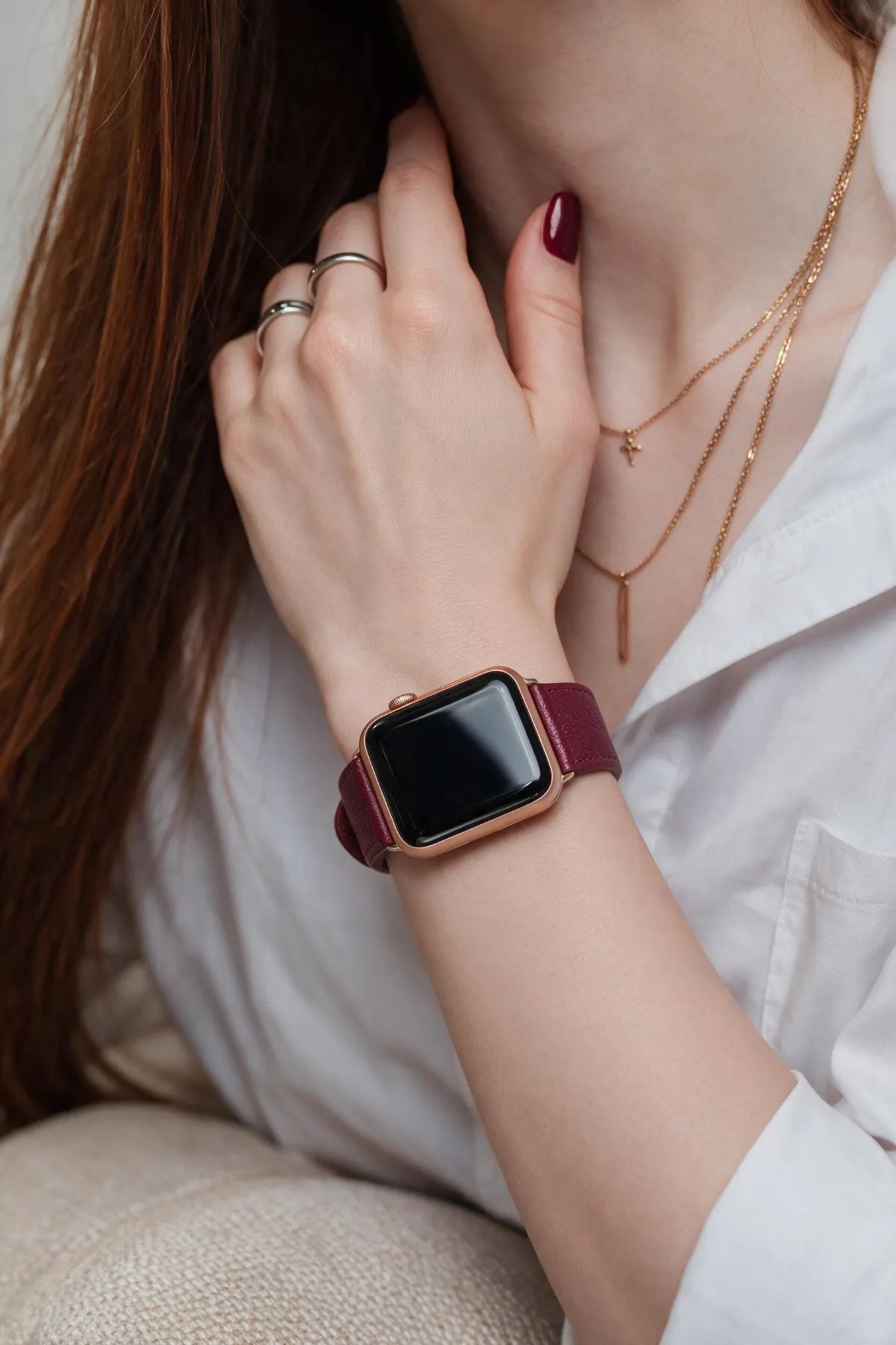 A close-up lifestyle shot of a woman wearing a smartwatch with a rose gold-toned case and a burgundy leather strap, styled with gold necklaces and rings.