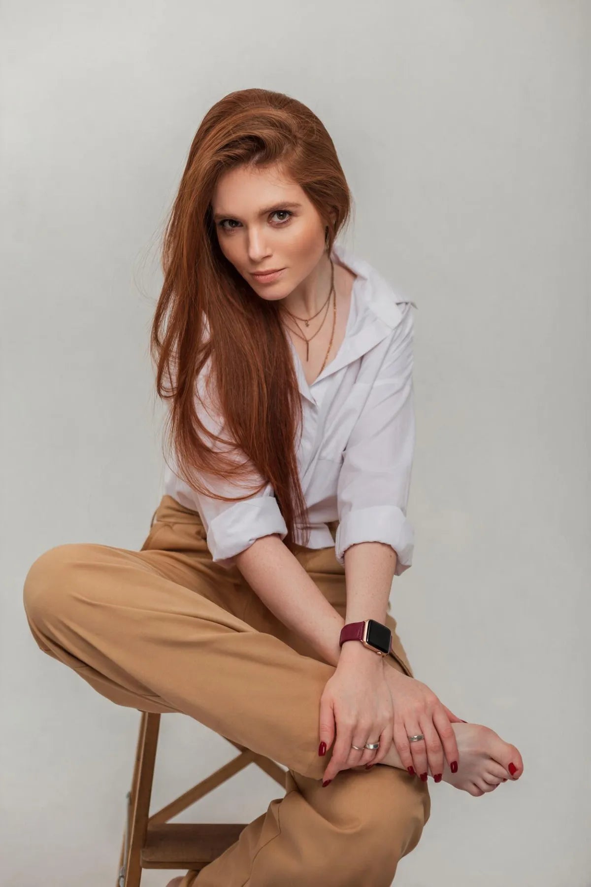 A woman with long red hair sits cross-legged on a wooden stool against a light gray studio background. She is wearing a white button-up shirt, tan pants, layered necklaces, a watch, and rings.