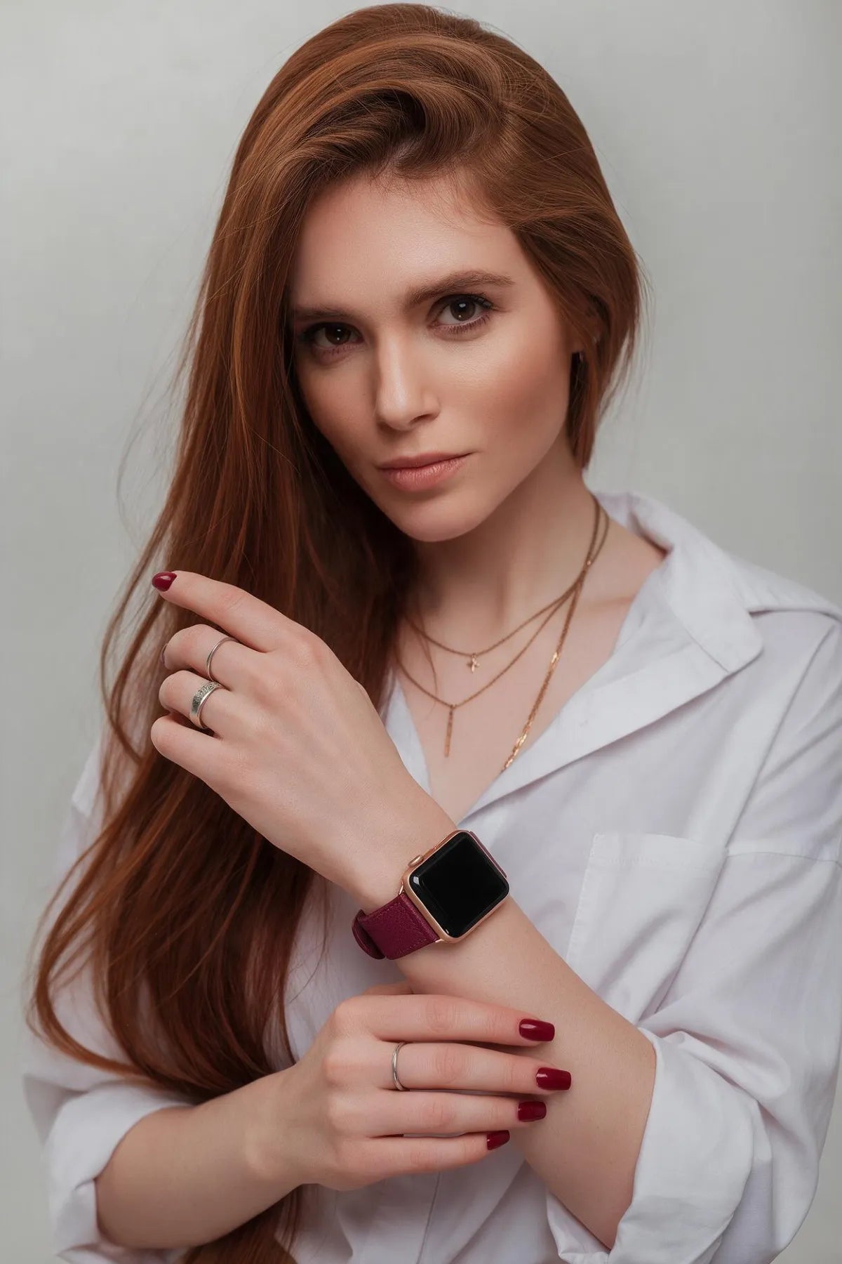 A woman with long red hair wearing a white shirt and gold necklaces, showcasing a smartwatch with a burgundy strap on her wrist against a light gray background.