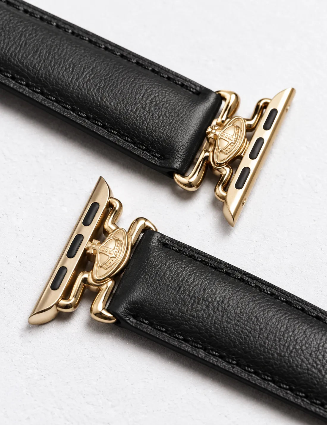 Close-up of a black leather watch strap with gold-toned hardware, showing the textured strap and a gold clasp with a crest emblem on a light gray background.