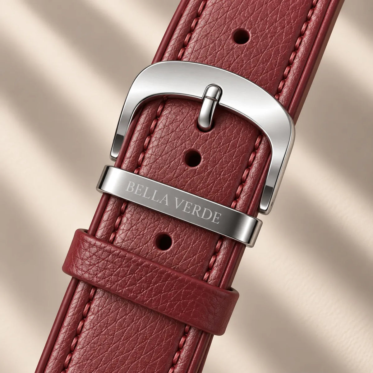 A close-up of a watch strap in textured burgundy leather with a polished silver-tone buckle. The metal keeper is engraved with the brand name 'BELLA VERDE'.