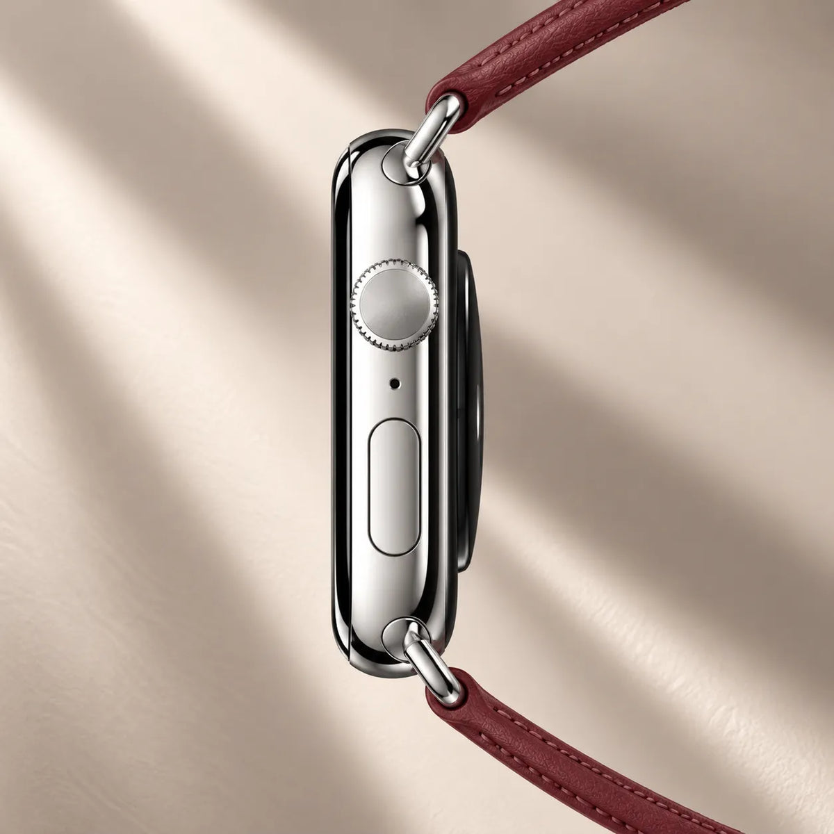 A close-up side view of a silver smartwatch with a burgundy leather strap, shown against a warm beige background with soft diagonal shadows.