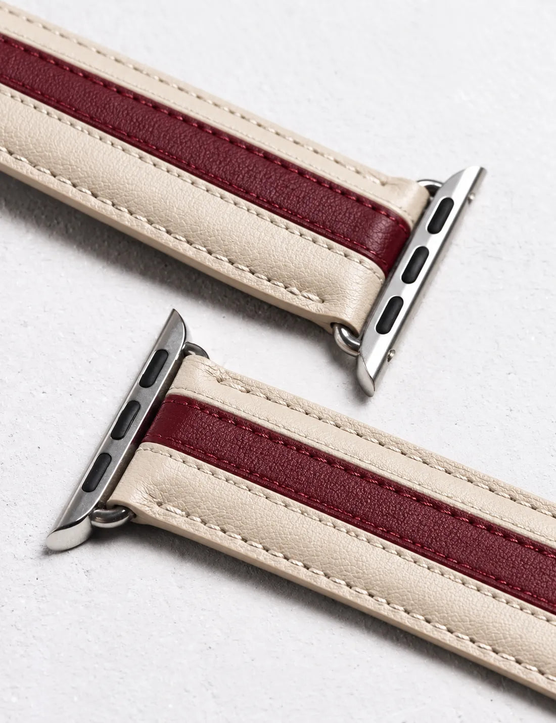 A close-up of a leather watch band with a cream-colored outer strap and a burgundy-red center stripe, shown with silver metal watch connectors on a light gray textured background.