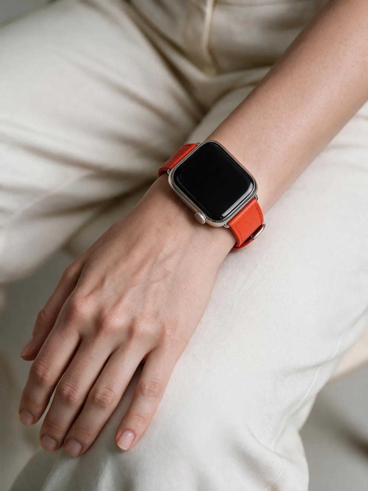 A close-up of a person's hand and wrist wearing a smartwatch with a bright orange leather band, dressed in light beige trousers.