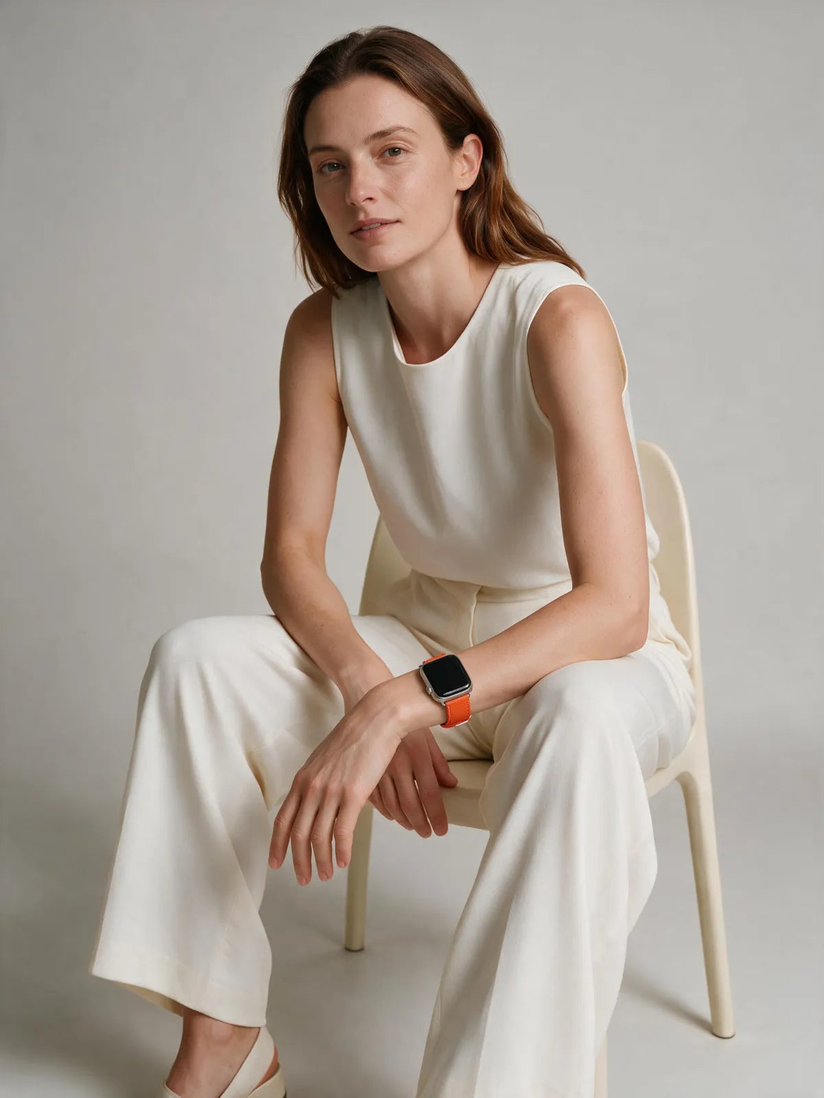 A woman sits on a light beige chair wearing a sleeveless cream top and matching wide-leg pants, with a smartwatch featuring an orange band on her wrist, against a neutral light gray background.