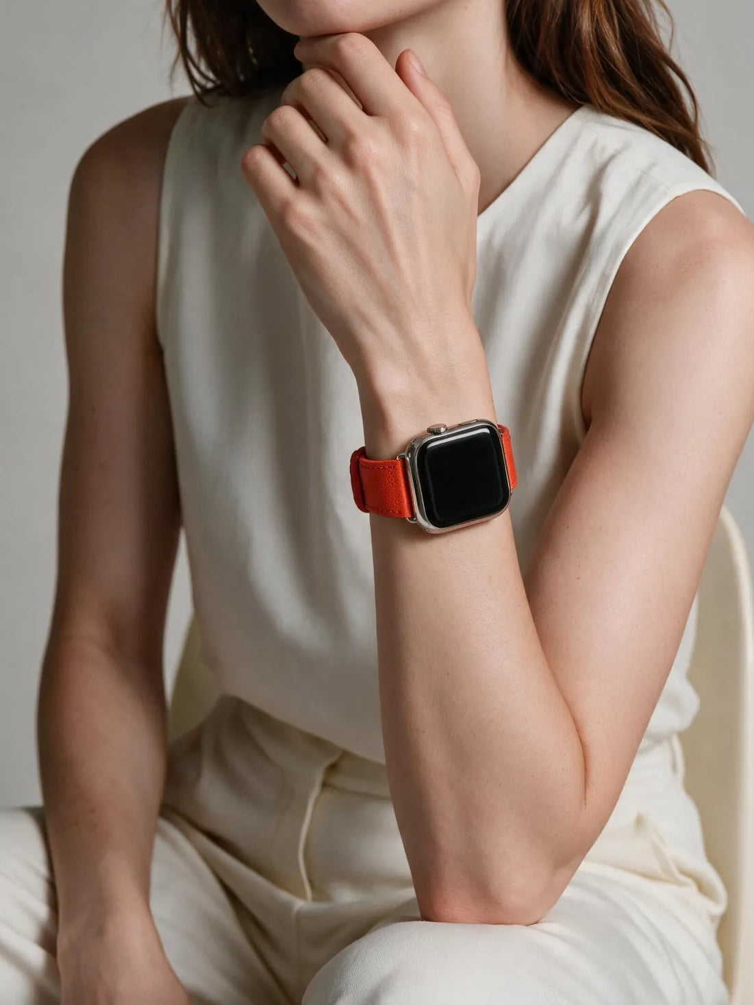 A woman wearing a smartwatch with a bright orange leather strap and a silver-toned metal case, seated against a neutral light gray background.