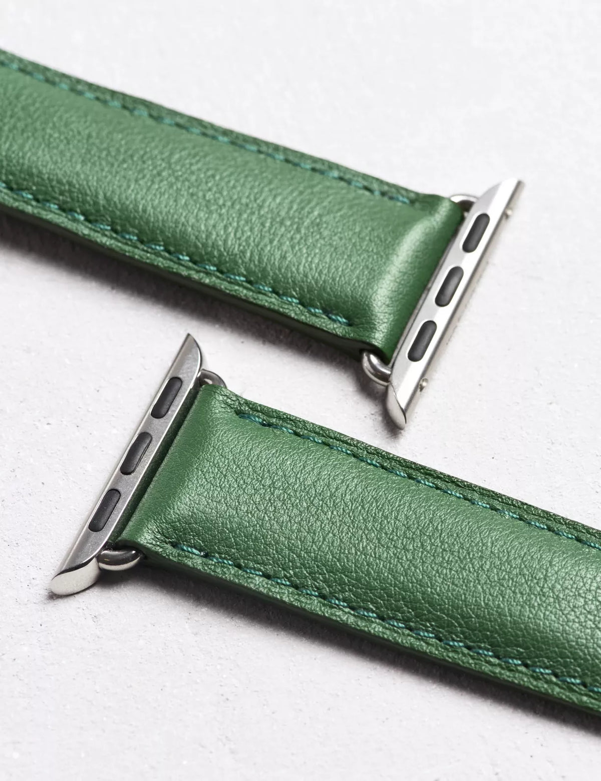 A close-up of a green leather watch band with silver metal hardware, laid diagonally on a light gray textured background.