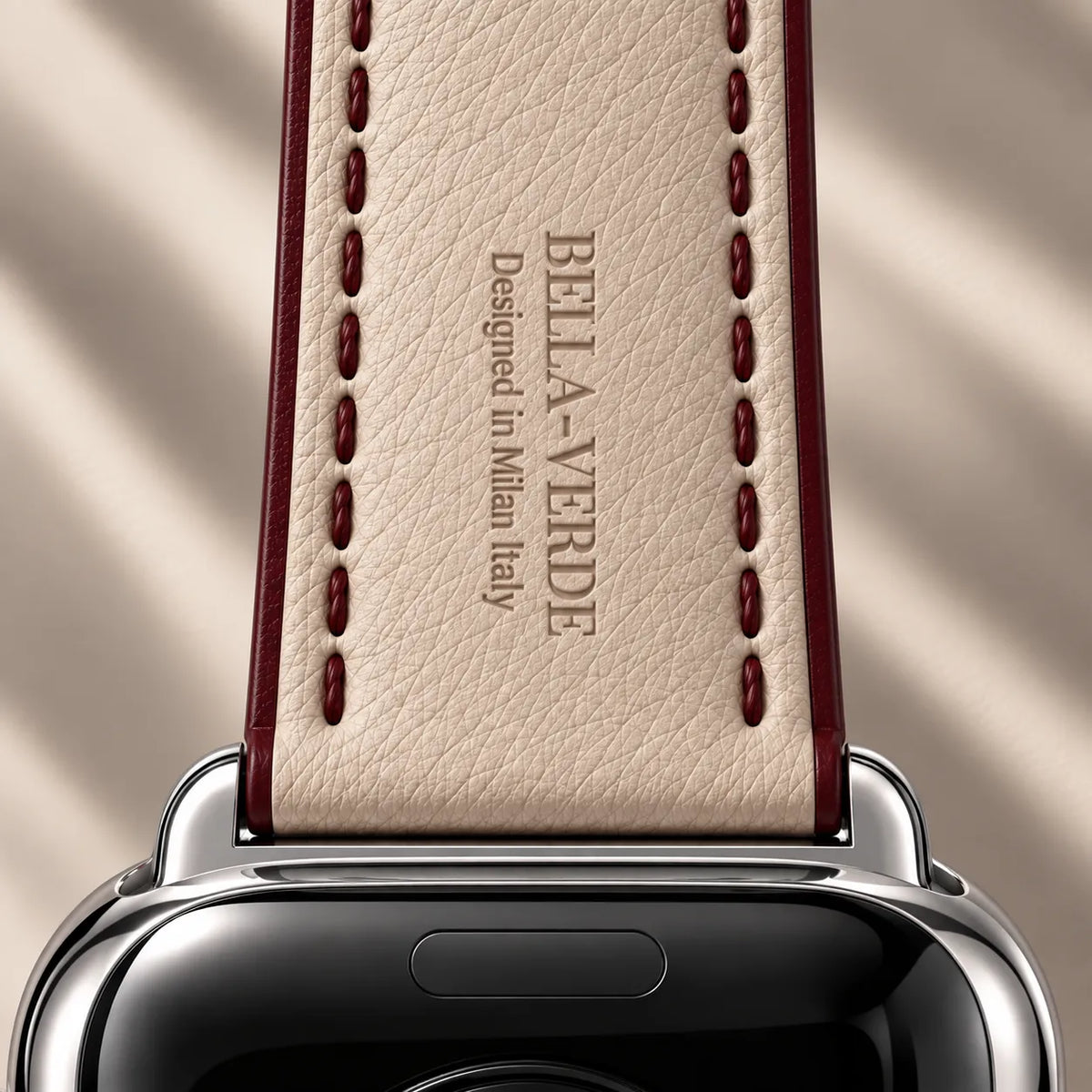Close-up of a smartwatch strap with a cream-colored textured leather inlay and dark red stitching, featuring the debossed brand name BELLA-VERDE on the back, attached to a silver watch case.
