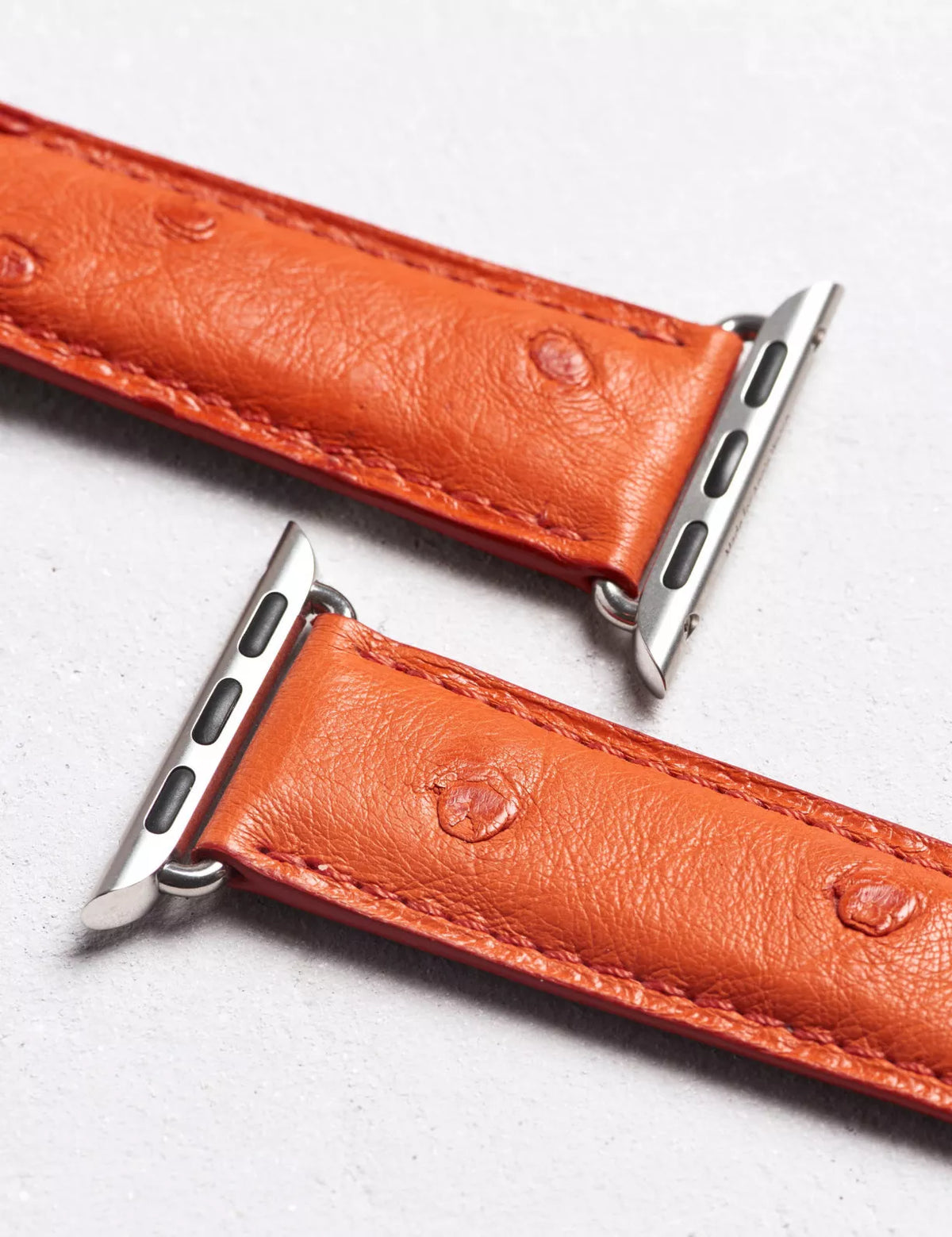 A close-up of an orange ostrich leather watch band with silver metal hardware, laid diagonally on a light gray textured surface.